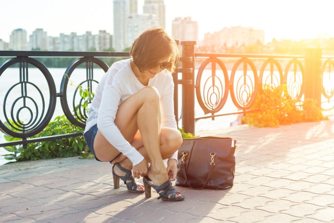 Outdoor summer portrait of mature woman in sunglasses with a bag and straightening shoes in the evening sunny city